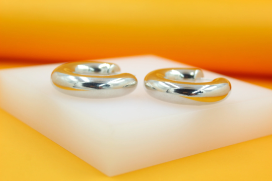 Two thick, polished silver hoop earrings rest on a white display block against an orange backdrop.