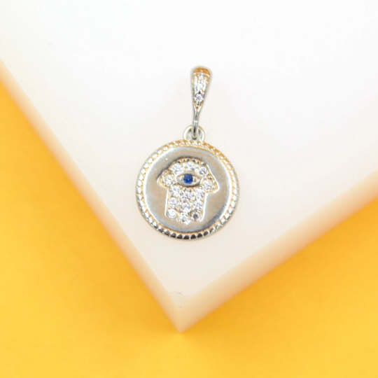 A silver Hamsa hand pendant featuring pavé-set cubic zirconia stones and a central blue evil eye rests against a white and yellow background.