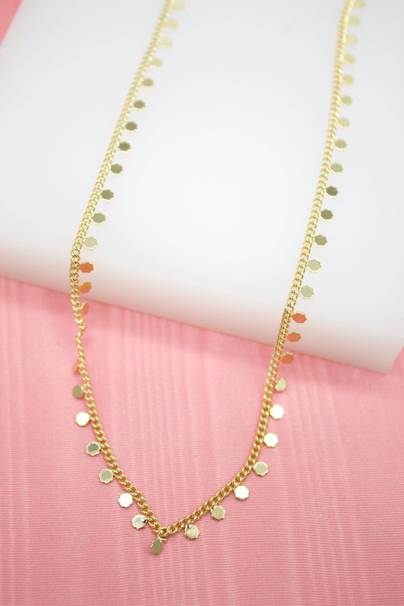 A delicate gold chain necklace featuring tiny dangling coin charms rests against a pink and white background.