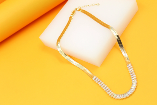 A gold herringbone chain necklace with a rhinestone-encrusted section rests on a white block against a vibrant yellow background.