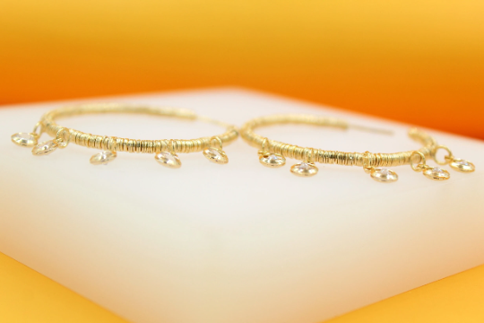 A pair of gold-wrapped hoop earrings with dangling crystal charms rests on a white display stand against a warm yellow background.