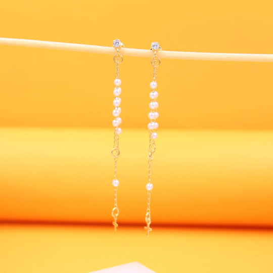 A pair of delicate gold chain earrings featuring dangling white pearls and tiny cross charms hang from a wooden stick against a bright yellow background.