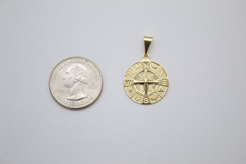 A gold compass pendant rests next to a US quarter coin for size comparison on a plain surface.
