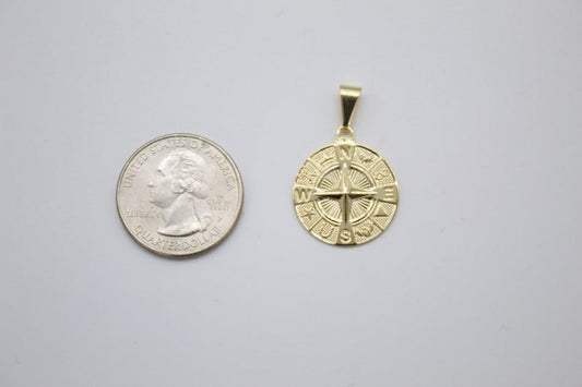 A gold compass pendant rests next to a US quarter coin for size comparison on a plain surface.