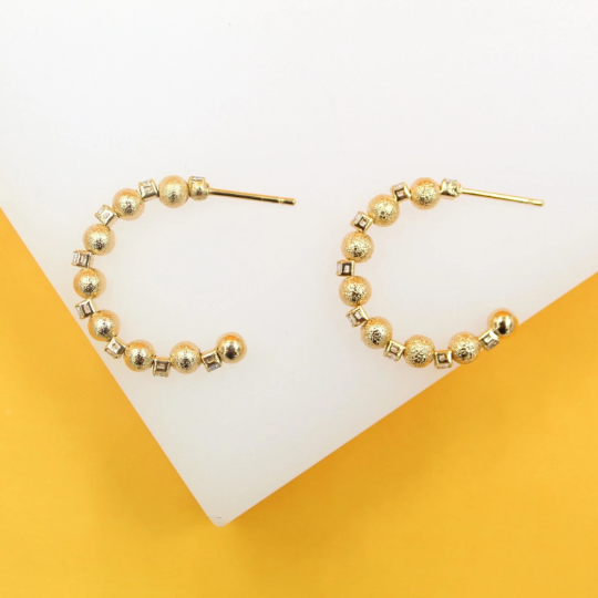 A pair of textured gold hoop earrings featuring alternating beaded spheres and tiny square gemstones rest on a white display surface against a vibrant yellow background.
