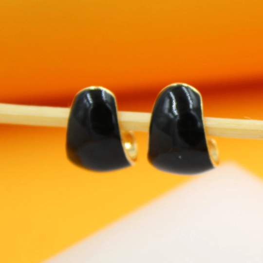 A pair of small, black enamel huggie hoop earrings rests delicately on a light wooden stick against an orange background.