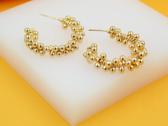 A pair of gold-plated hoop earrings embellished with tiny beaded details rests on a white display block against an orange backdrop.