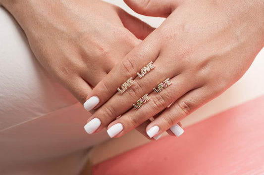 A woman displays her hands with white manicured nails while wearing four gold "LOVE" cocktail rings encrusted with sparkling stones.
