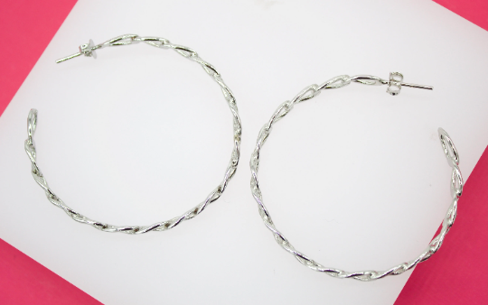 A pair of large silver twisted hoop earrings rests on a white display surface against a vibrant pink background.