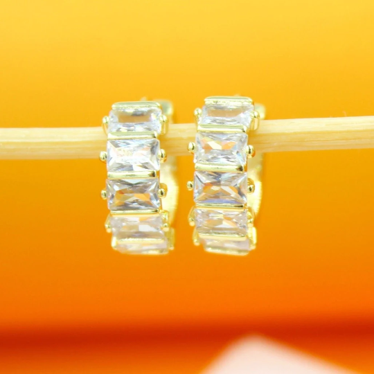 Two gold hoop earrings featuring sparkling baguette-cut cubic zirconia stones rest on a wooden stick against an orange background.