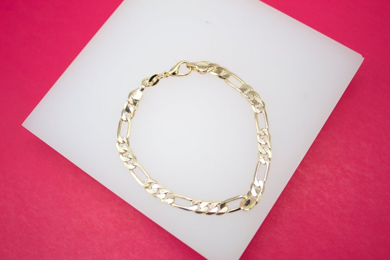 A shiny gold Figaro chain bracelet rests on a white display stand against a vibrant pink background.
