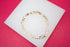 A shiny gold Figaro chain bracelet rests on a white display stand against a vibrant pink background.