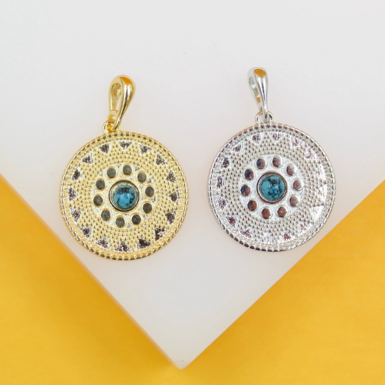 A gold pendant and a silver pendant featuring intricate patterned rims and central turquoise stones rest side by side on a white display surface.