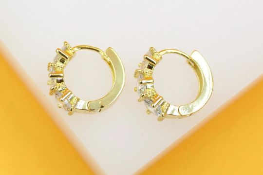 A pair of gold huggie hoop earrings embedded with sparkling cubic zirconia stones rests against a light surface.