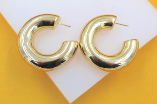 A pair of thick, polished gold-plated hoop earrings rests on a white display stand against a vibrant yellow background.