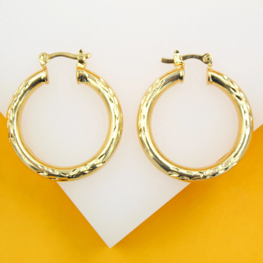 A pair of polished gold hoop earrings with delicate textured detailing rests against a contrasting white and yellow background.