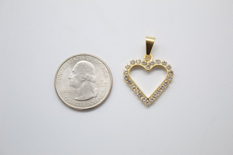 A gold heart-shaped pendant studded with sparkling rhinestones rests next to a US quarter coin for a size comparison on a plain background.