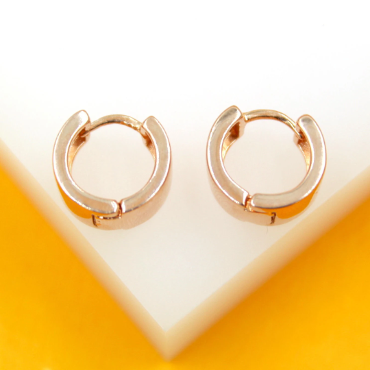 A pair of polished rose gold huggie hoop earrings rests against a white and yellow background.