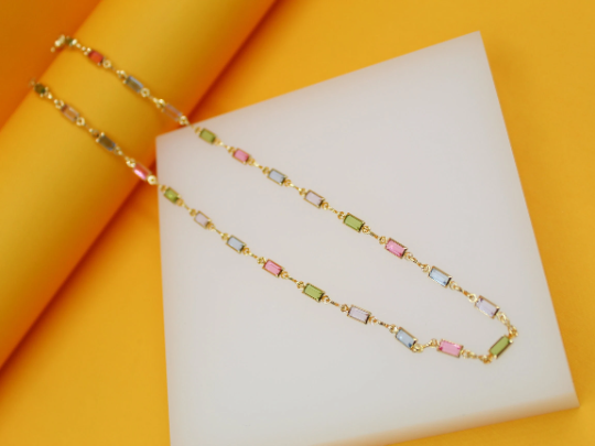 A gold chain necklace featuring colorful rectangular crystals rests gracefully on a white display block against a vibrant yellow background.