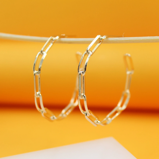 Two gold paperclip chain hoop earrings hang side by side against a vibrant orange background.