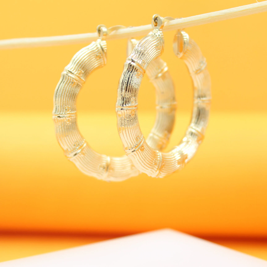 A pair of textured gold bamboo hoop earrings hang delicately from a wooden stick against a warm yellow background.