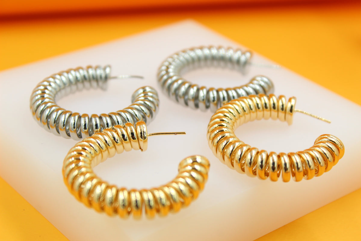Two pairs of chunky, spiral hoop earrings in silver and gold rest on a white display block against an orange backdrop.