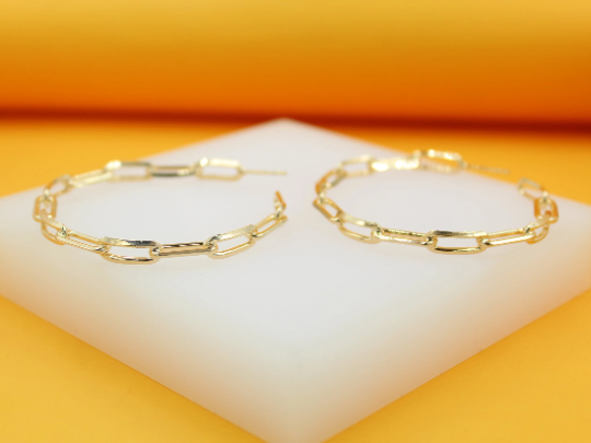 A pair of gold paperclip chain hoop earrings rests on a white display block against an orange backdrop.