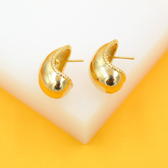 A pair of glossy gold dome hoop earrings featuring beaded trim rests on a white and yellow display stand.