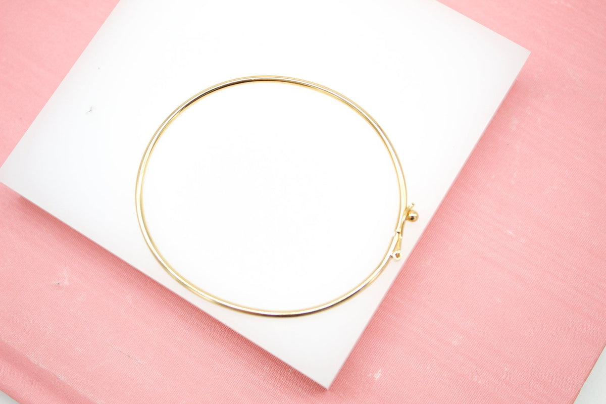 A sleek gold hoop bracelet rests on a white display against a soft pink background.