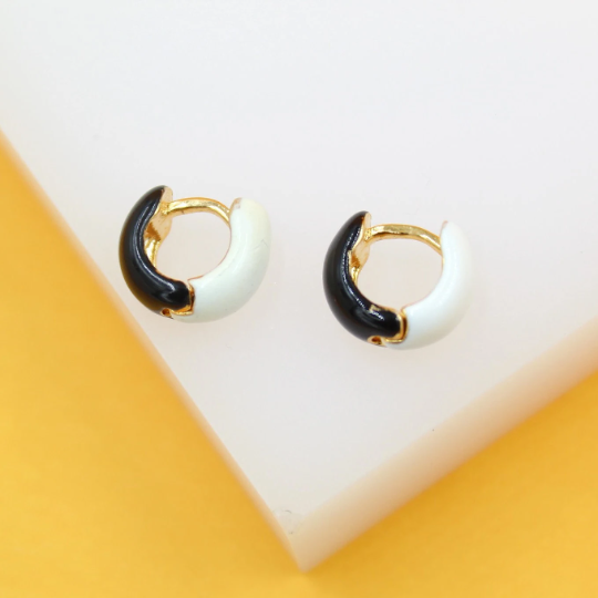 A pair of gold huggie hoop earrings featuring contrasting black and white enamel details rests on a white display surface.
