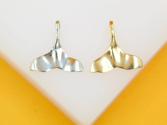 Two whale tail charms in silver and gold rest side by side on a clean white surface.