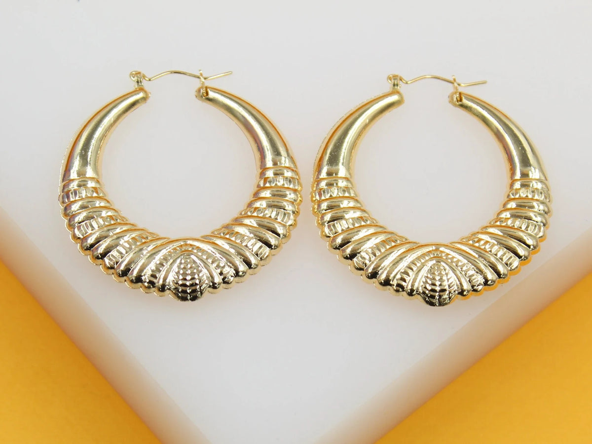 A pair of chunky gold hoop earrings with intricate textured details rest on a white display stand.