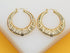 A pair of chunky gold hoop earrings with intricate textured details rest on a white display stand.