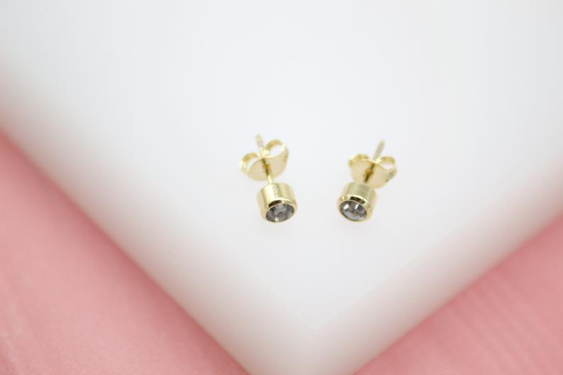 A pair of gold stud earrings featuring small, bezel-set cubic zirconia stones rests on a white display surface.