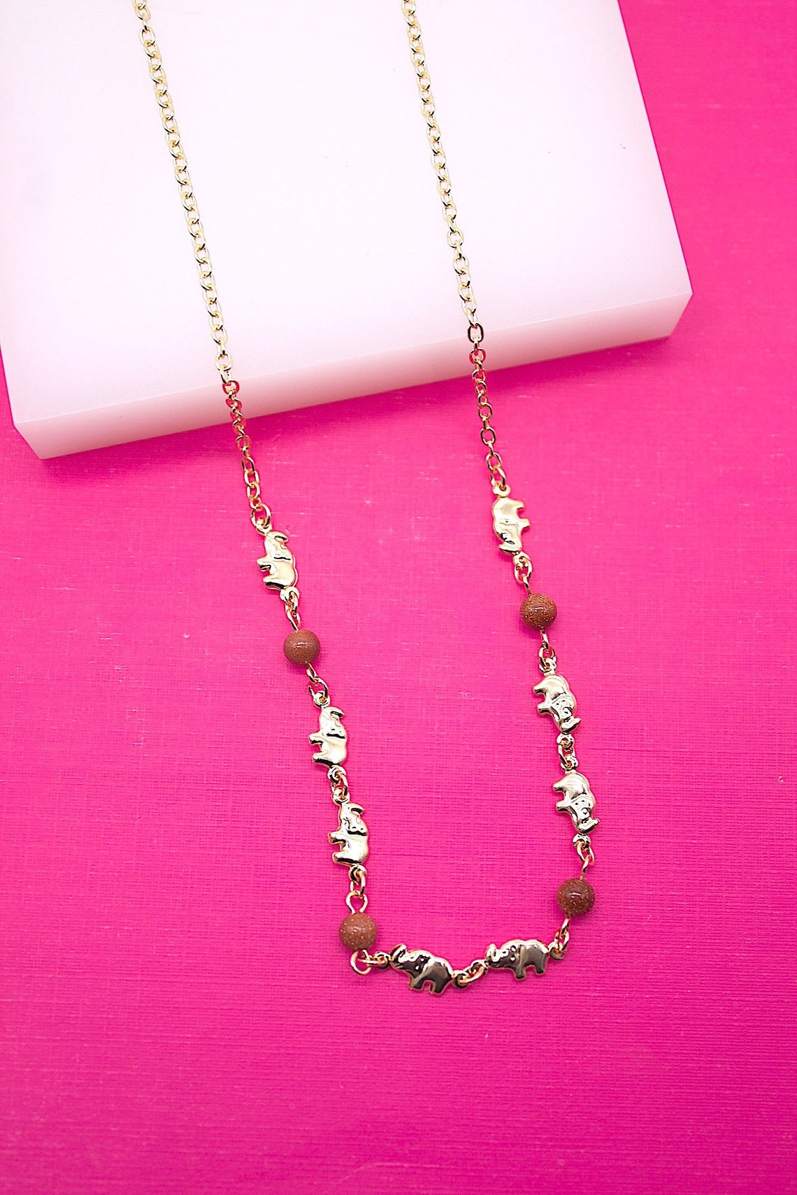 A gold-link necklace featuring alternating elephant charms and brown gemstone beads rests against a vibrant pink and white background.