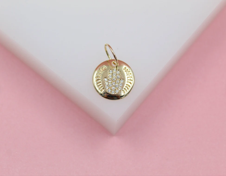 A gold hamsa pendant featuring pave diamonds rests on a white and pink display surface.