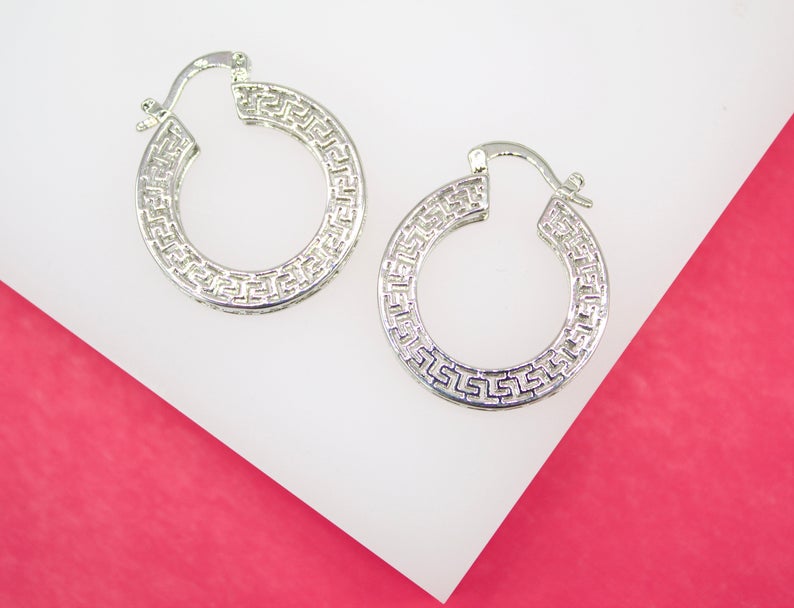 A pair of silver hoop earrings featuring an intricate Greek key pattern rests on a white and pink background.