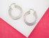 A pair of silver hoop earrings featuring an intricate Greek key pattern rests on a white and pink background.