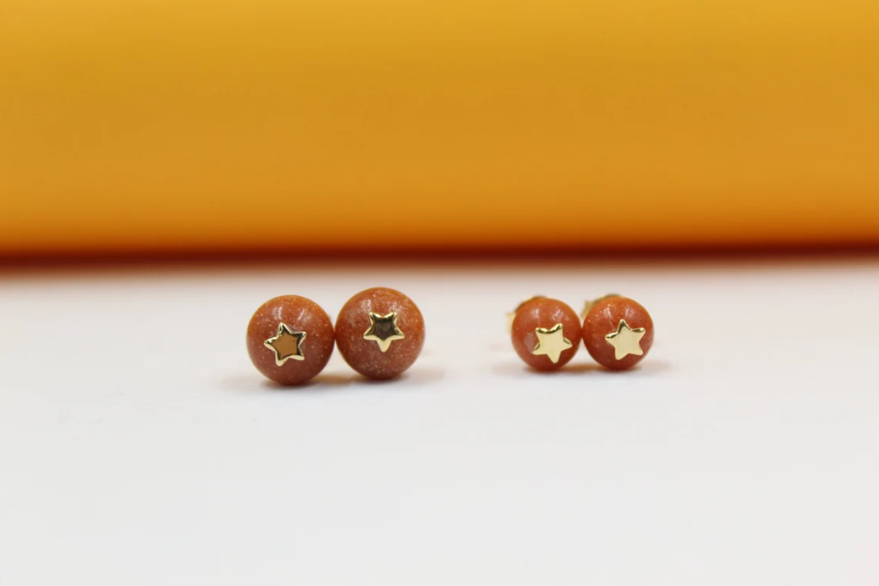 Two pairs of orange sandstone ball stud earrings feature delicate gold star charms against a bright yellow and white backdrop.