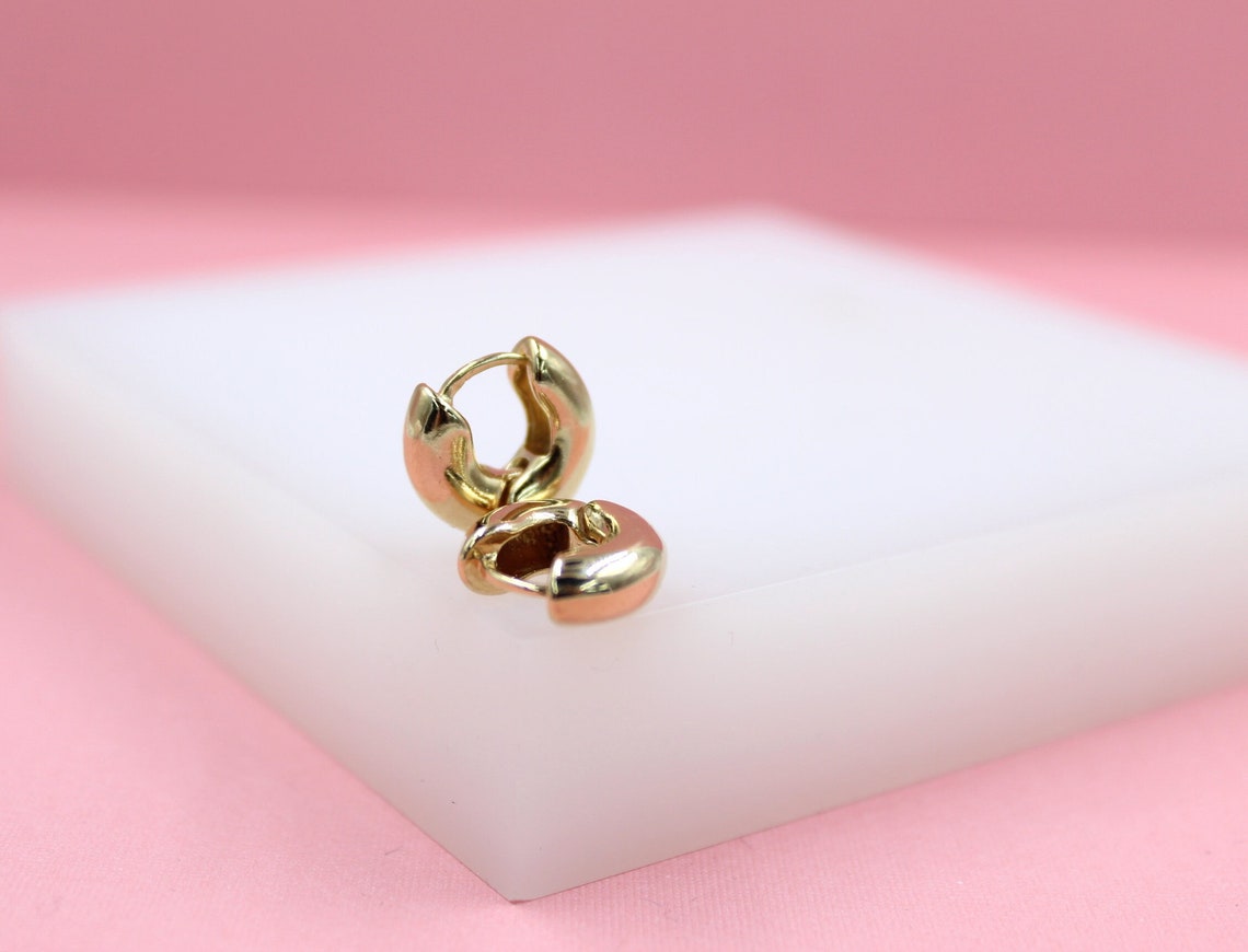 A pair of polished gold huggie hoop earrings rests on a white display block against a soft pink backdrop.