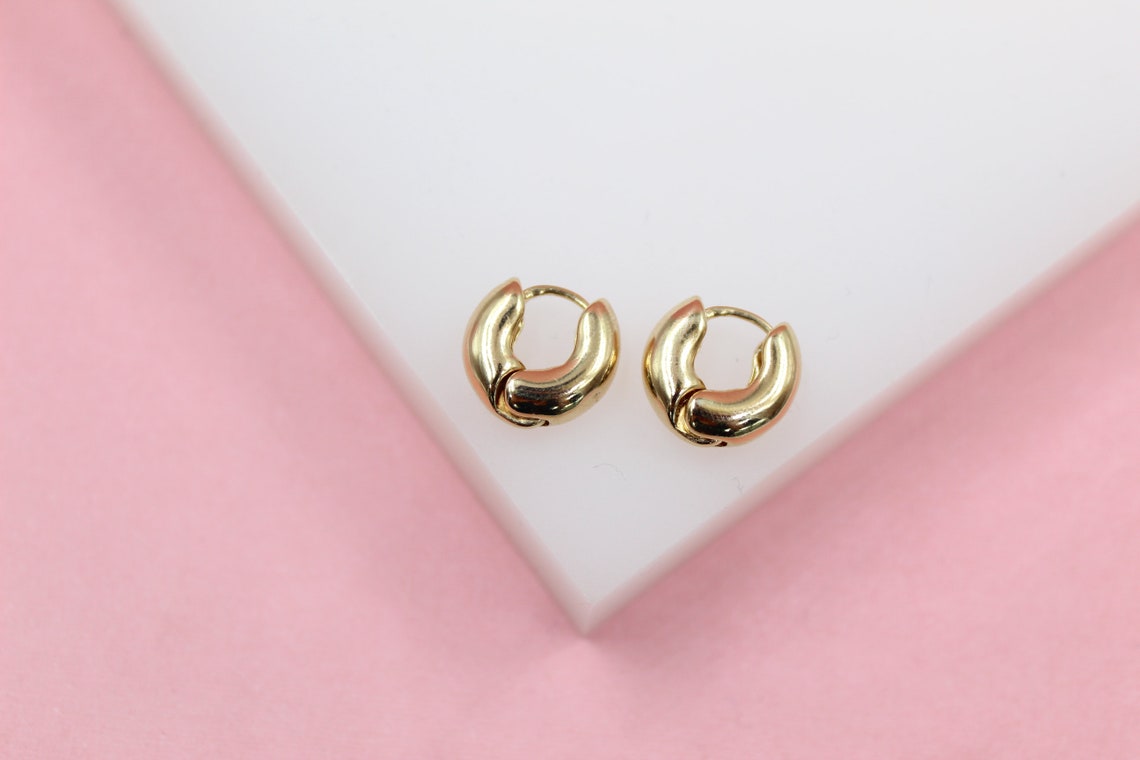 A pair of small, chunky gold huggie hoop earrings rests on a white display against a pink background.