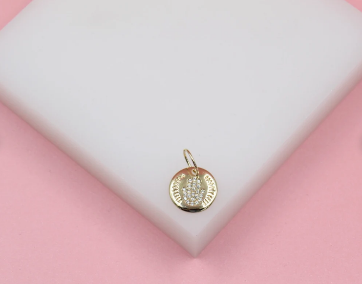 A gold Hamsa hand pendant featuring sparkling pave diamonds rests on a white display surface against a pink background.