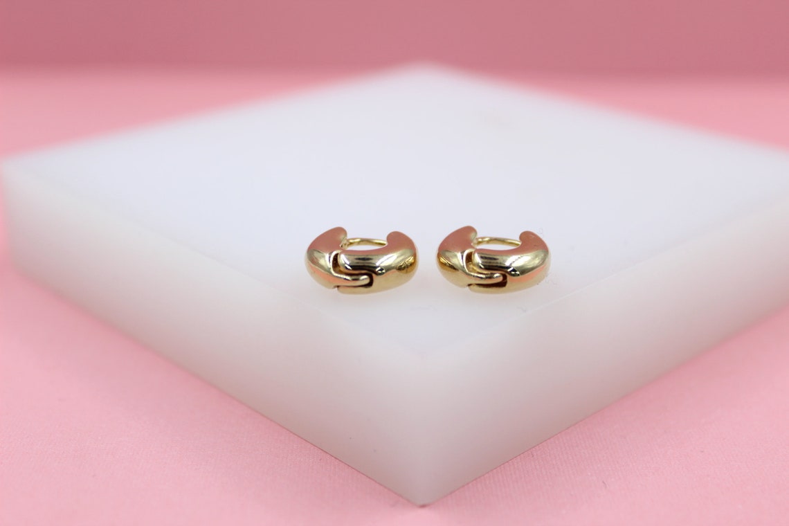 A pair of polished gold huggie hoop earrings rests on a white block against a soft pink backdrop.