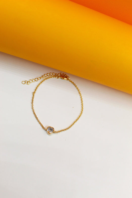 A delicate gold chain bracelet features a sparkling heart-shaped cubic zirconia pendant against a bright yellow and white background.