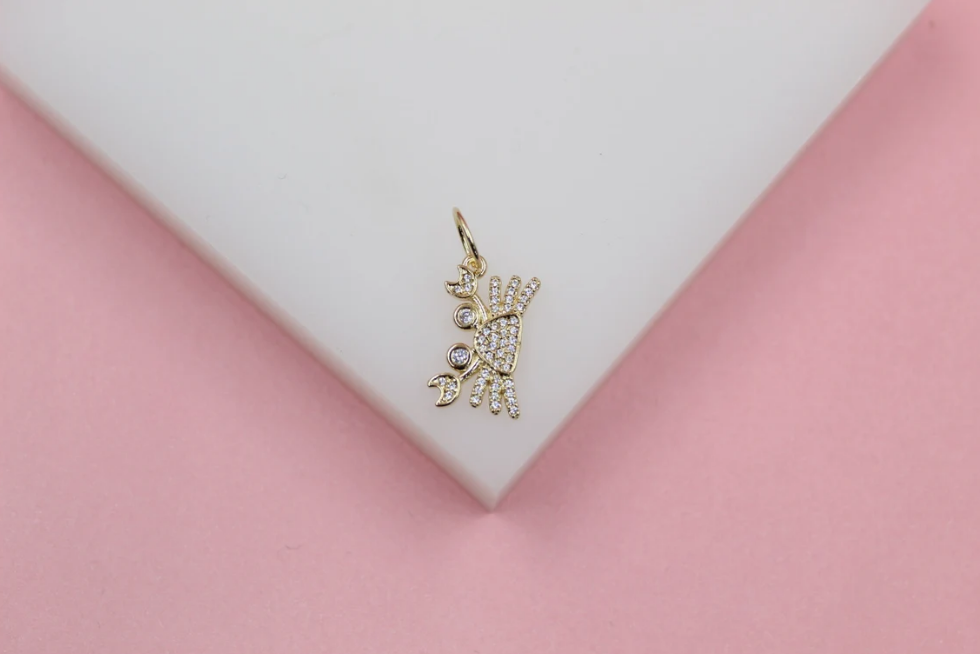 A gold crab charm encrusted with sparkling cubic zirconia stones rests on a white display surface against a pink background.