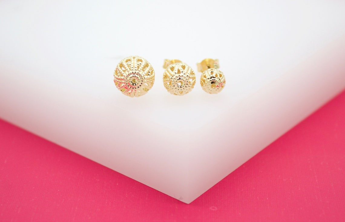 Three delicate gold filigree ball stud earrings display intricate openwork patterns on a white and pink surface.