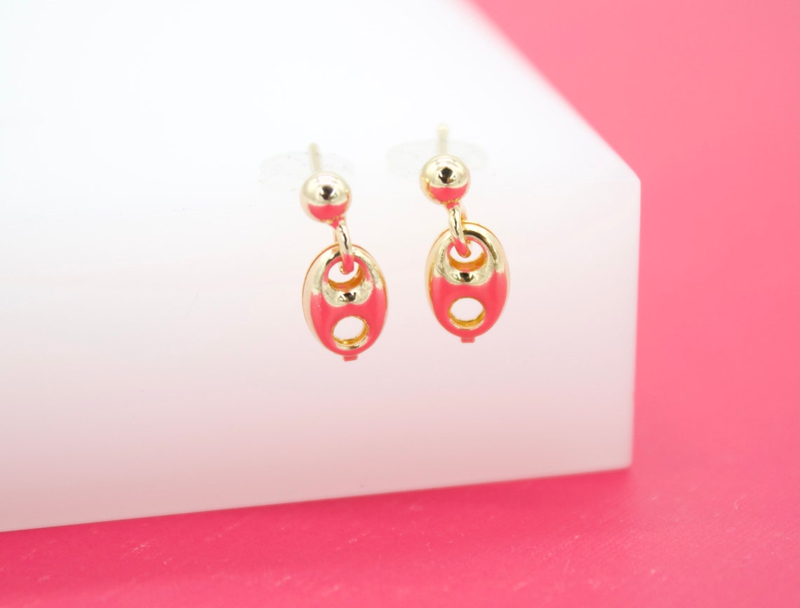 A pair of gold stud earrings featuring vibrant pink enamel dangling charms rests on a white display block against a pink background.