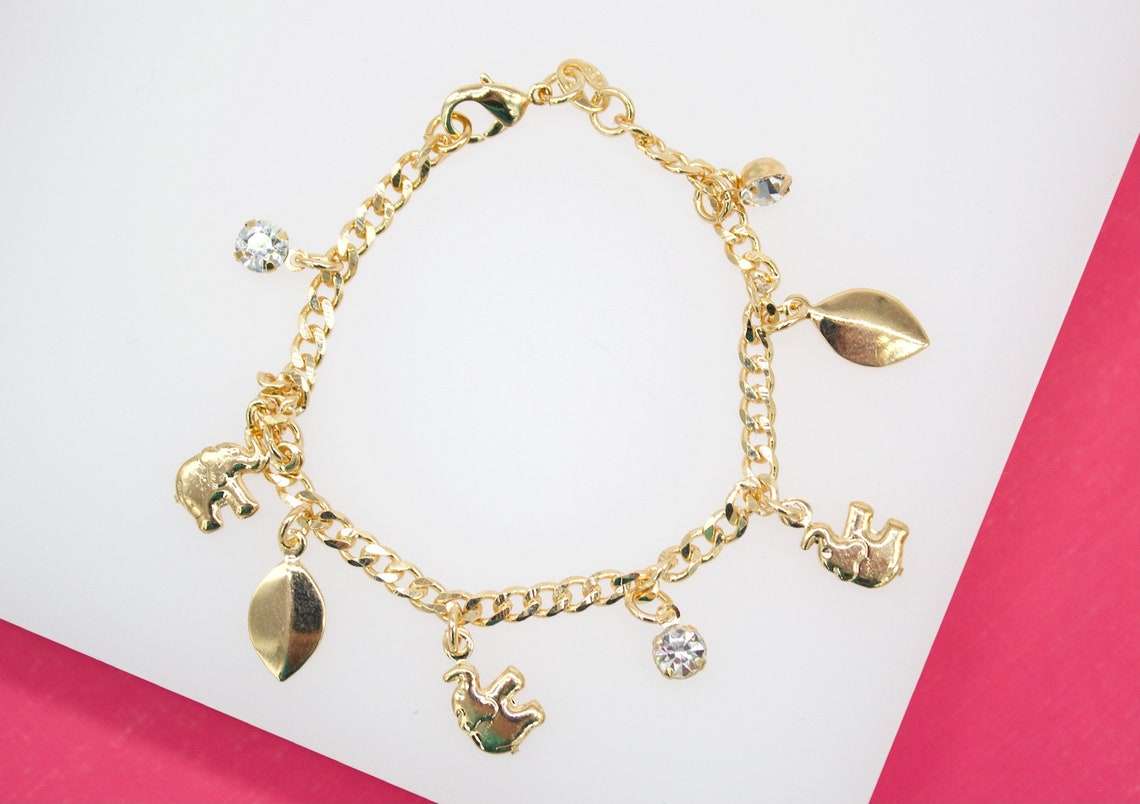 A gold chain charm bracelet features dangling elephant, leaf, and sparkling rhinestone charms against a white and pink background.