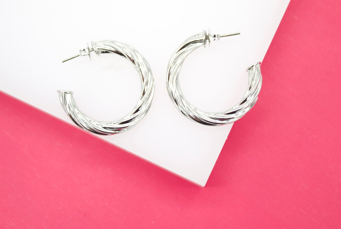 A pair of polished silver twist hoop earrings rests on a white display surface against a vibrant pink background.