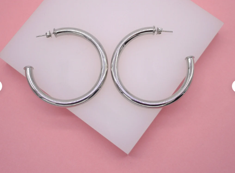 A pair of polished silver chunky hoop earrings rests on a white display pad against a pink background.